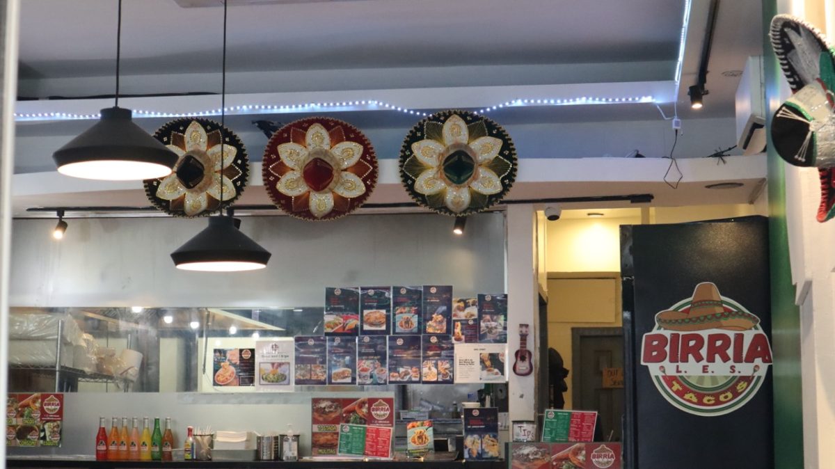 The interior of a Mexican restaurant contains decorative sombreros hanging on the wall above the kitchen counter, with a large sign on the right features the words "BIRRIA L.E.S. TACOS”.