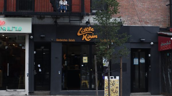 A storefront with a black awning that reads “Little Kirin Sandwiches Salads”. A sign shows various photos of menu items outfront.