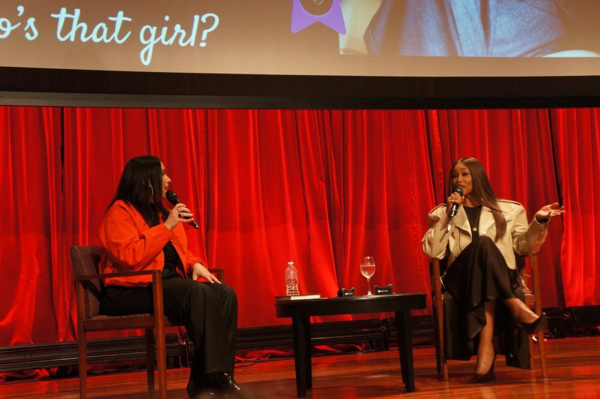 Two women sit on a stage, speaking into microphones in front of a red backdrop.