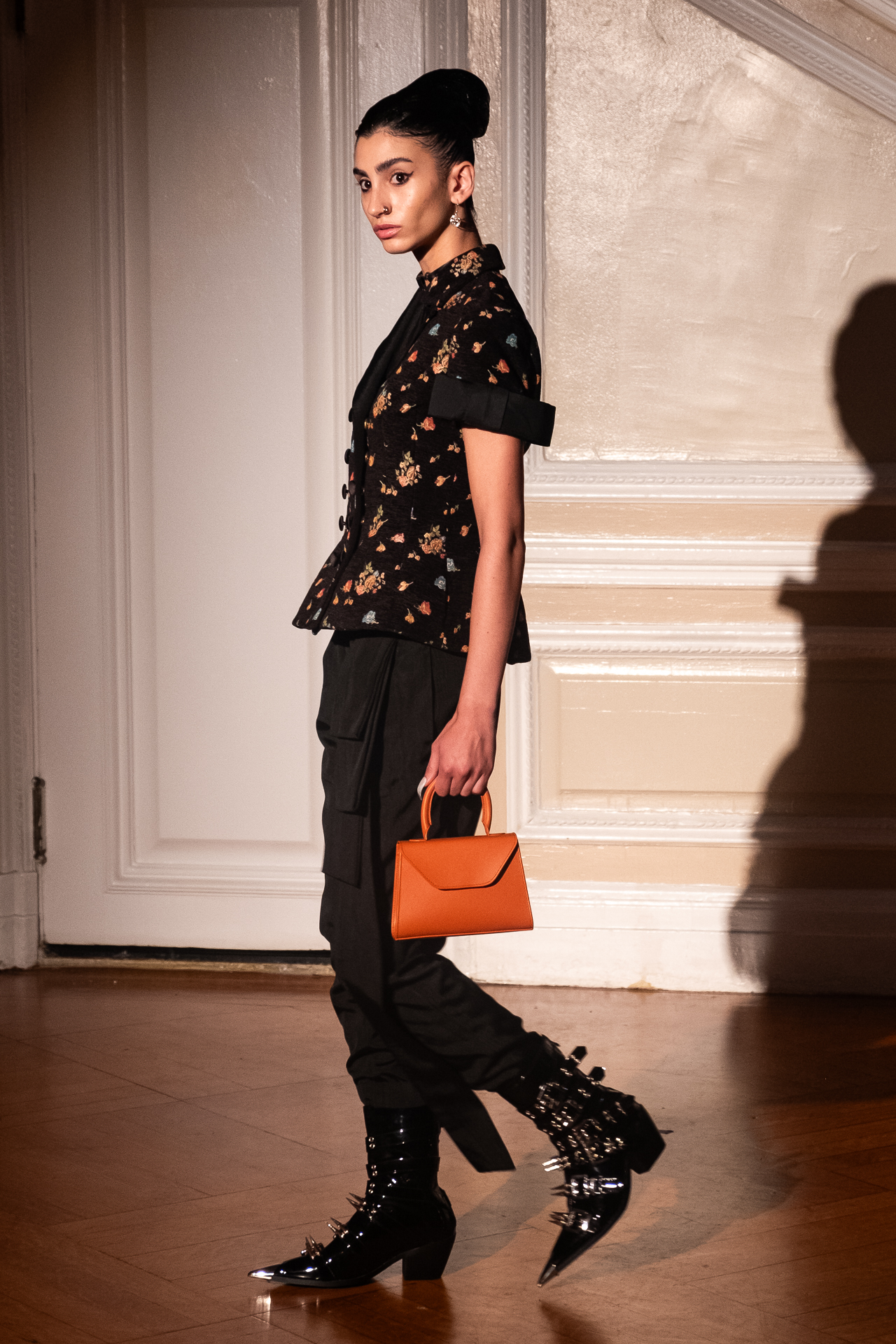 A model mid-walk wearing a high-collared, black, floral, short-sleeve button-down shirt with a bow on the sleeve, an ankle-length, fitted black bottom with a layered detail hanging from the waist and spiky black pointed-toe ankle boots. The model holds a small tan handbag.