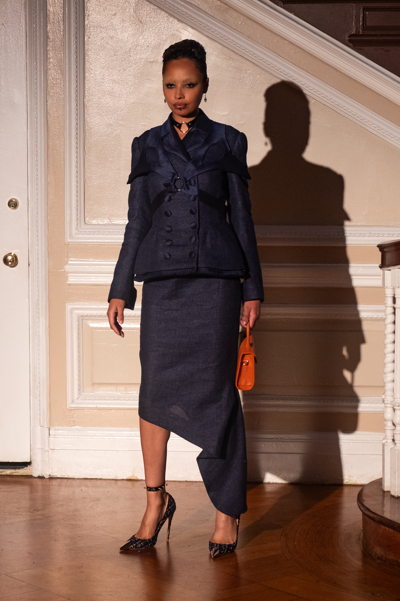 A model stands in front of a wall wearing a double-breasted peplum jacket with long sleeves and a draped skirt with an asymmetrical hem in matching shiny blue fabric. The model holds a small tan handbag and wears pointed blue heels with ankle straps.