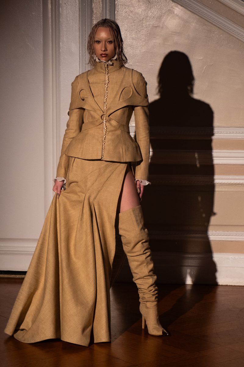 A model standing in front of a flashlight, wearing a high khaki-beige collar with layered circular cut-out on two sides, with a side slit skirt and knee high boots.