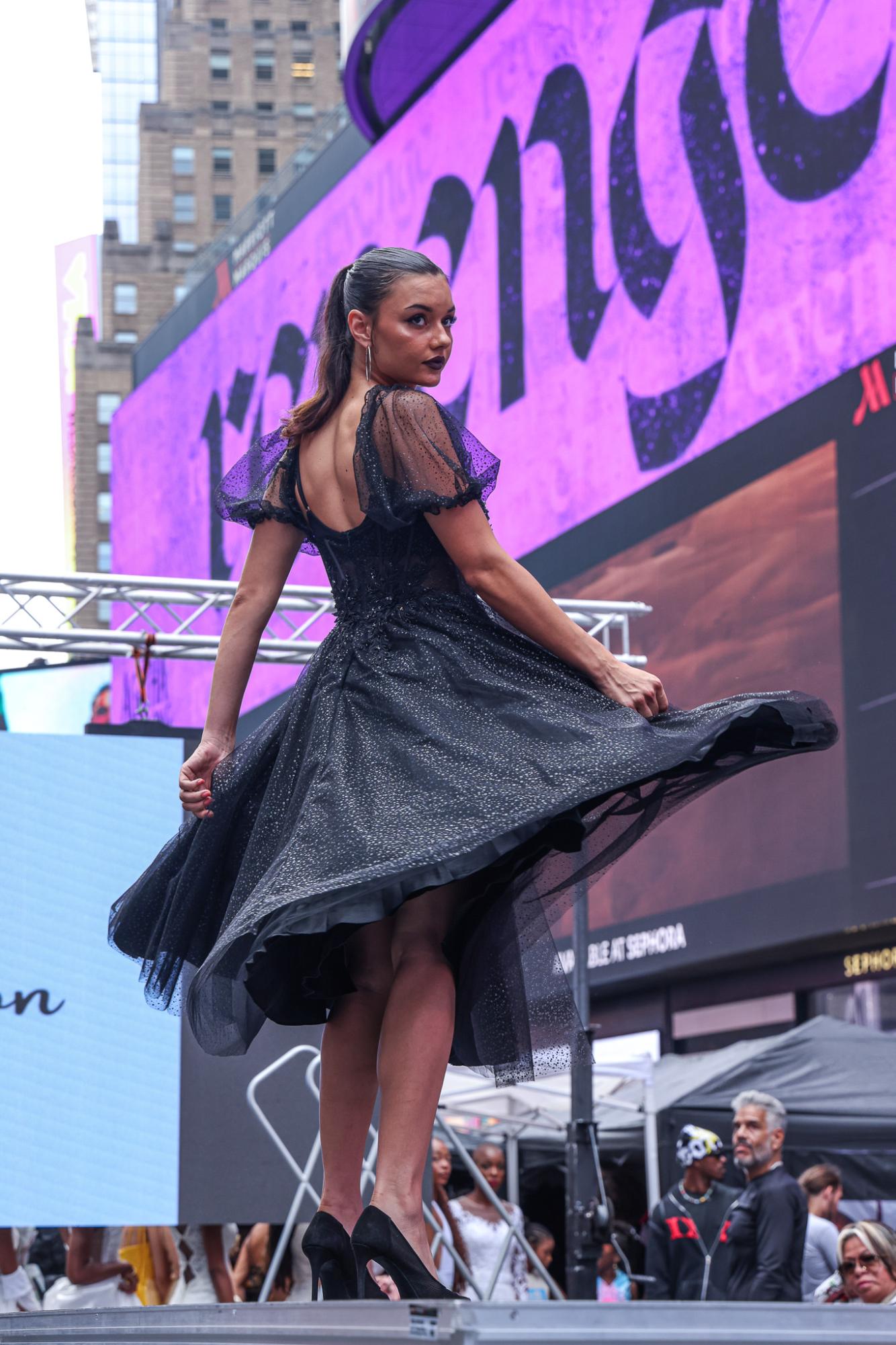 A model in a black sparkly dress with mesh shoulders and black heels spins on a runway.