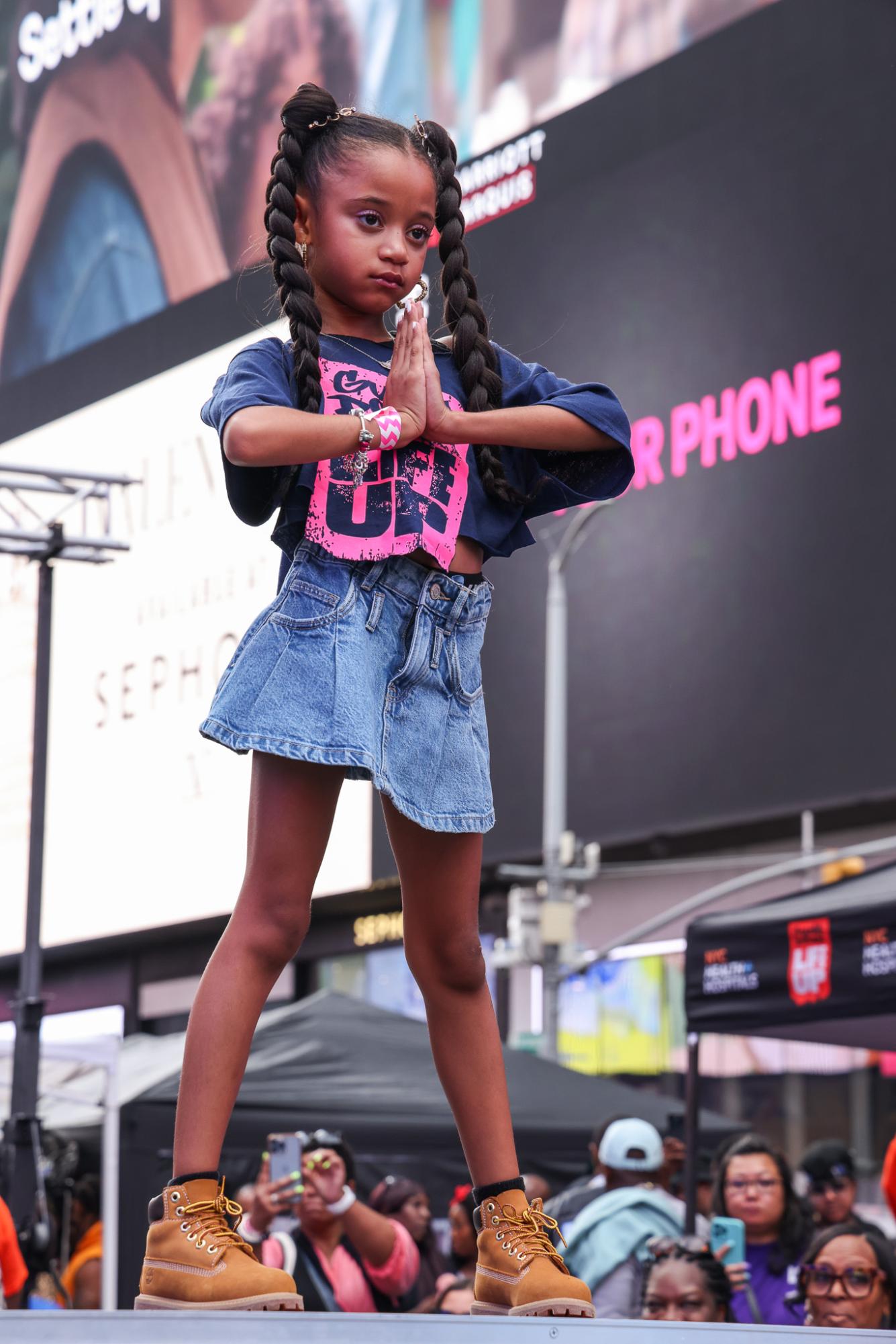 A young girl poses on the runway in a blue and pink shirt with “GUNS DOWN, LIFE UP” written on it and a denim skirt.