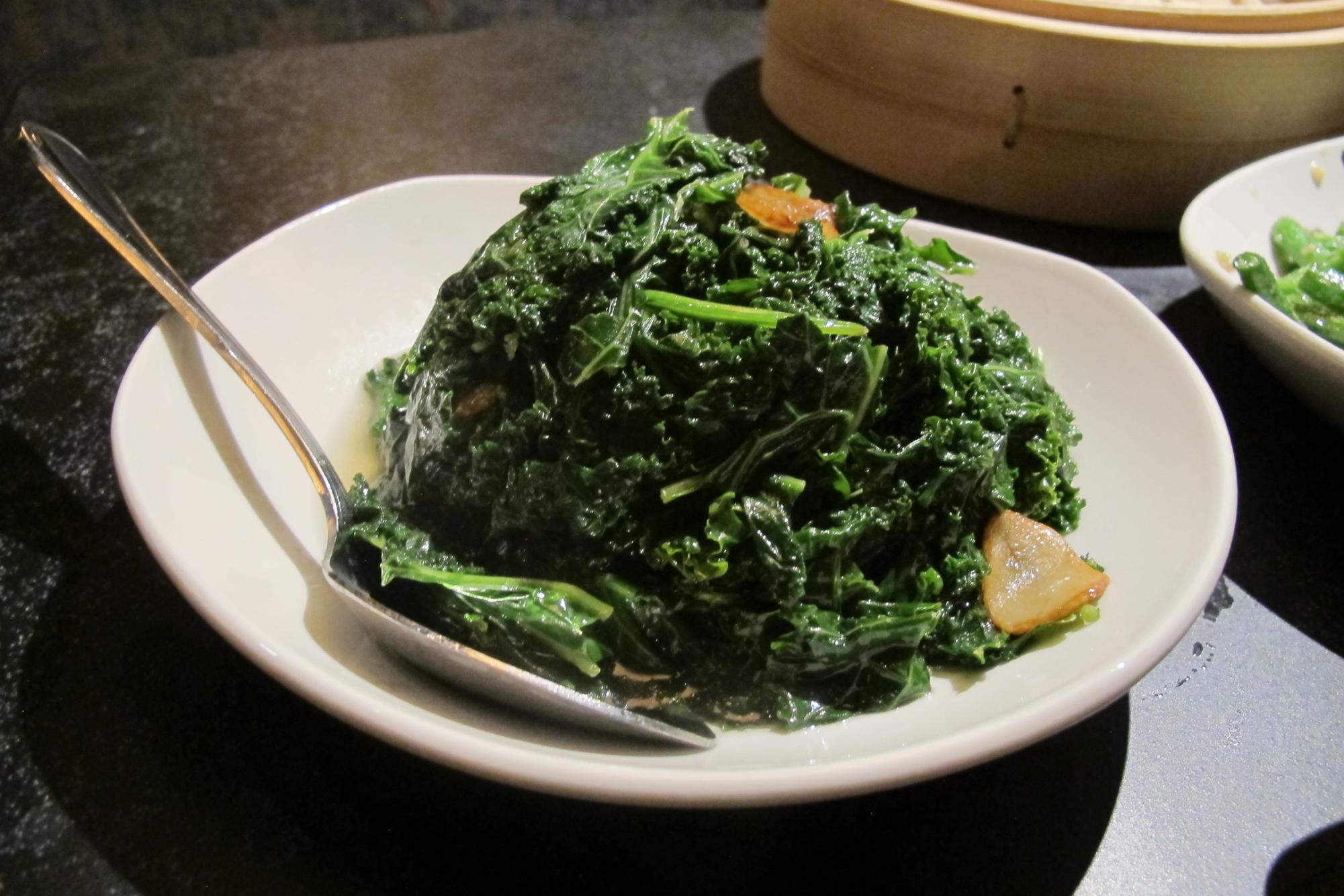 Kale with garlic slices in a white bowl, with a metal spoon resting on one side.