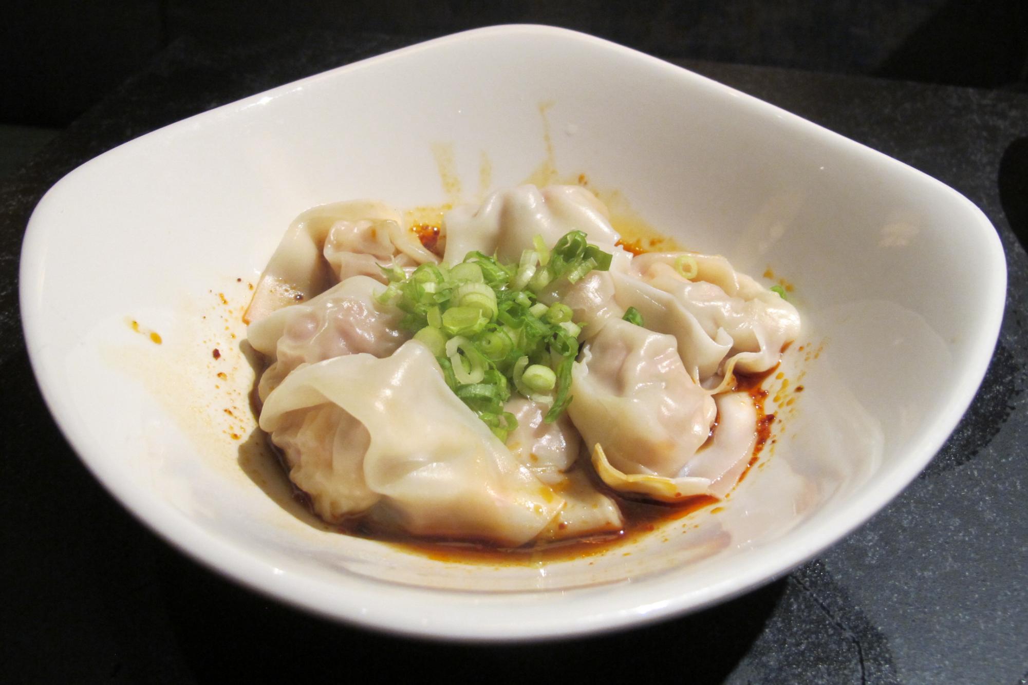 Shrimp and kurobuta pork spicy wontons sitting on chili oil in a white bowl, sprinkled with scallions.