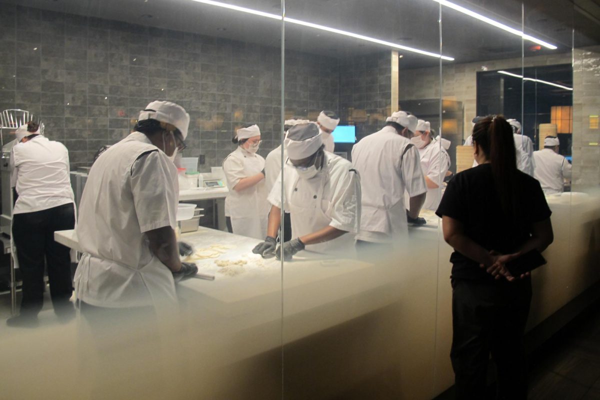 A woman in black watches chefs preparing food in the kitchen behind a glass wall.