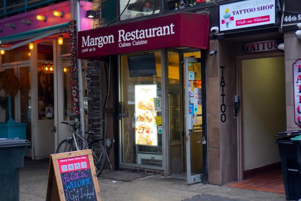 A storefront with a red awning that reads, “Margon Restaurant Cuban Cuisine” in white lettering with a brightly lit menu in the window and the door propped open.