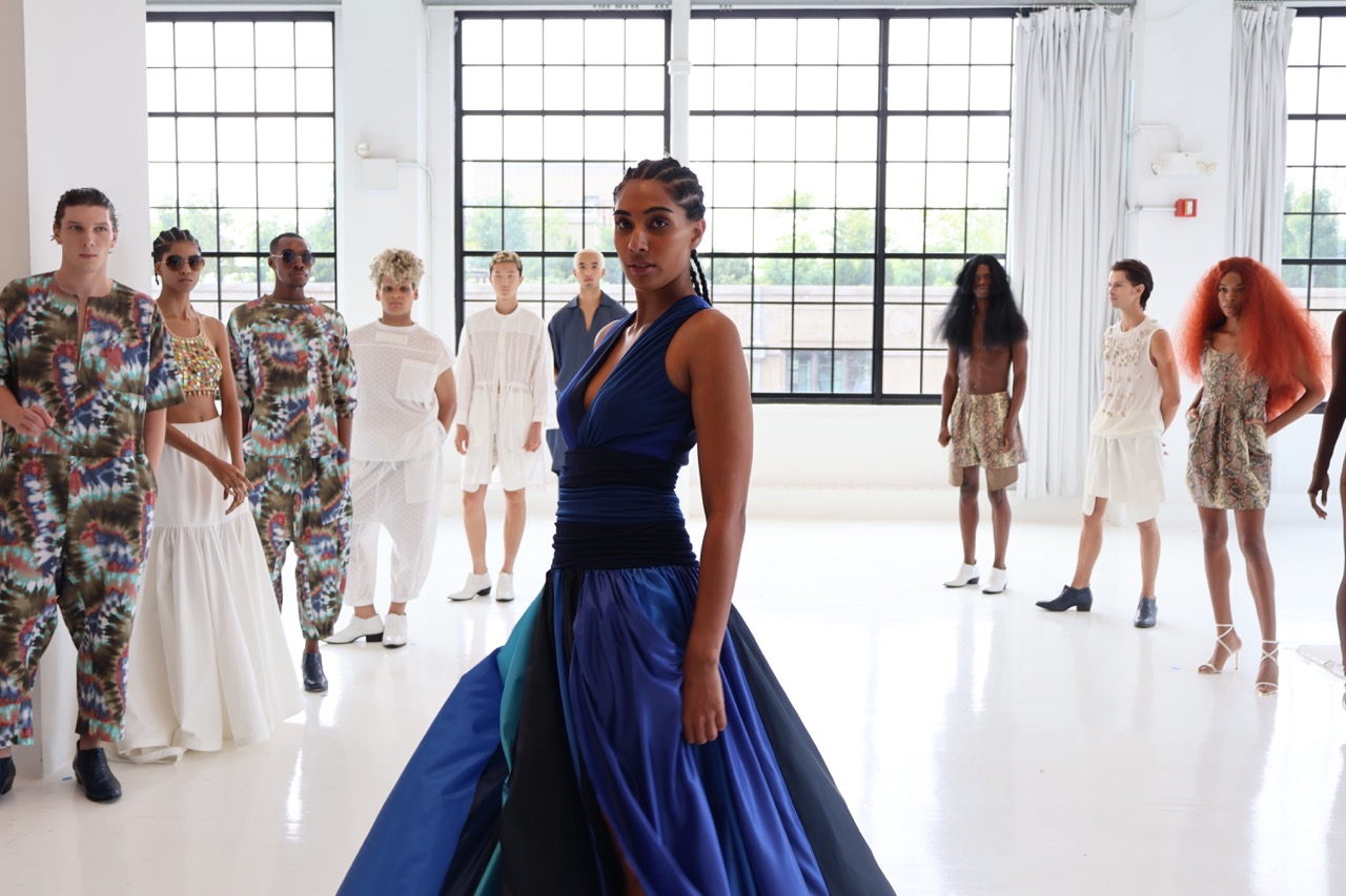 A model stands in the center of a white room wearing a long, flowing royal blue dress with a low neckline. The skirt of the dress includes multiple shades of blue. Nine other models stand behind her.