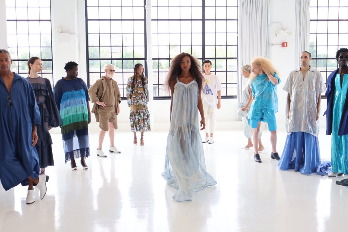 A model wearing a long light-blue spaghetti-strap dress walks in the middle of 10 other models standing in a circle.