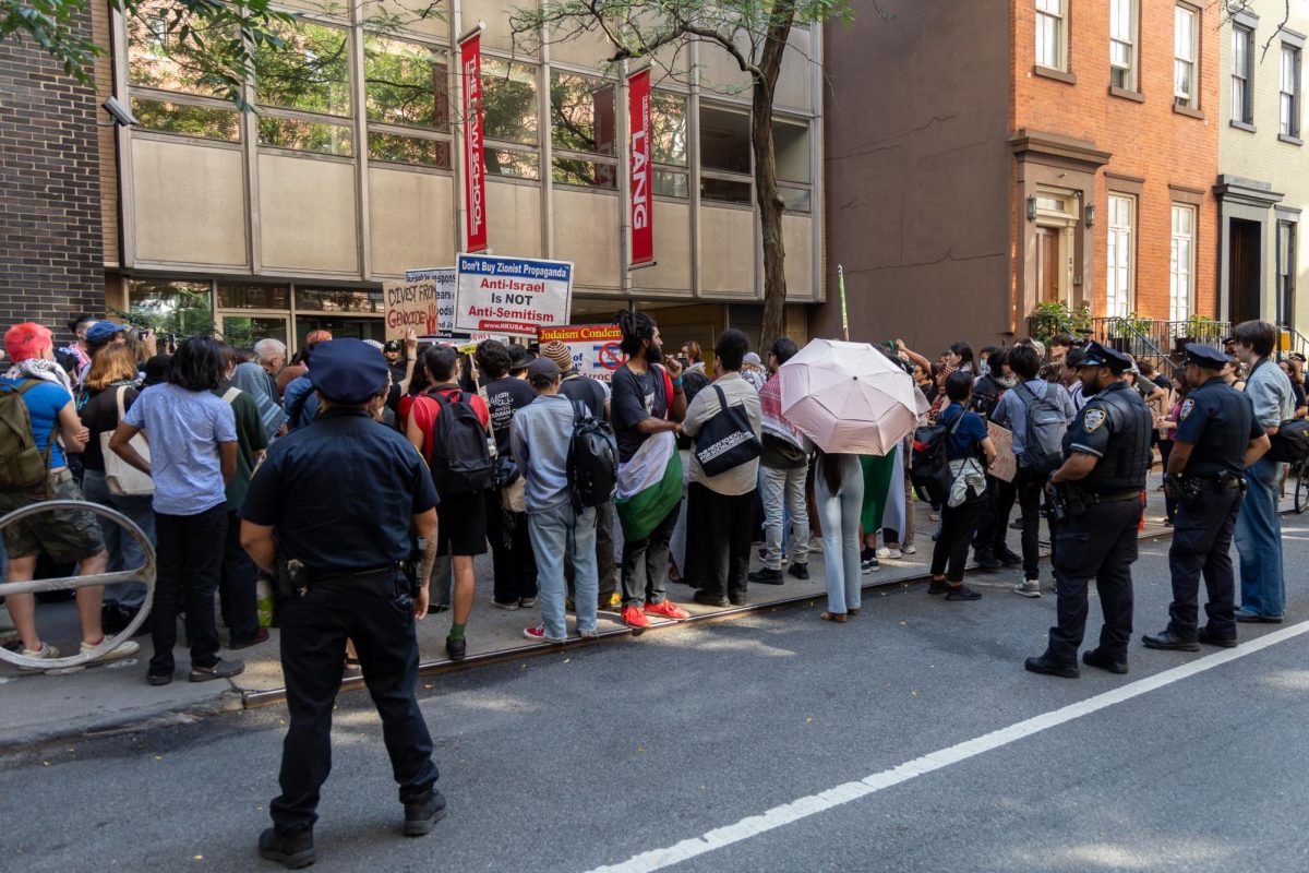 A large group of people stand on a sidewalk with signs reading, “Don’t Buy Zionist Propaganda. Anti-Israel is NOT Anti-Semitism” and Palestinian flags. Three police officers stand overlooking the protesters.