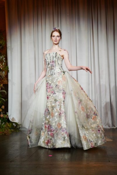 A model in a dress with a corset top and tulle skirt, with pink, purple and white florals all over it. She’s wearing a flower crown and has floral accents on her arm.