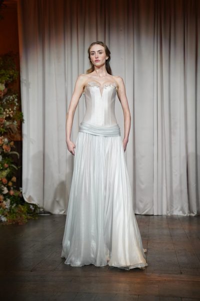 A woman in a silky, all white gown. The gown has a corset top and there are delicate details of white flowers at her chest line.