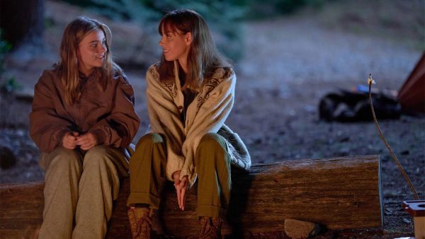 Two women sit on a log bench smiling.