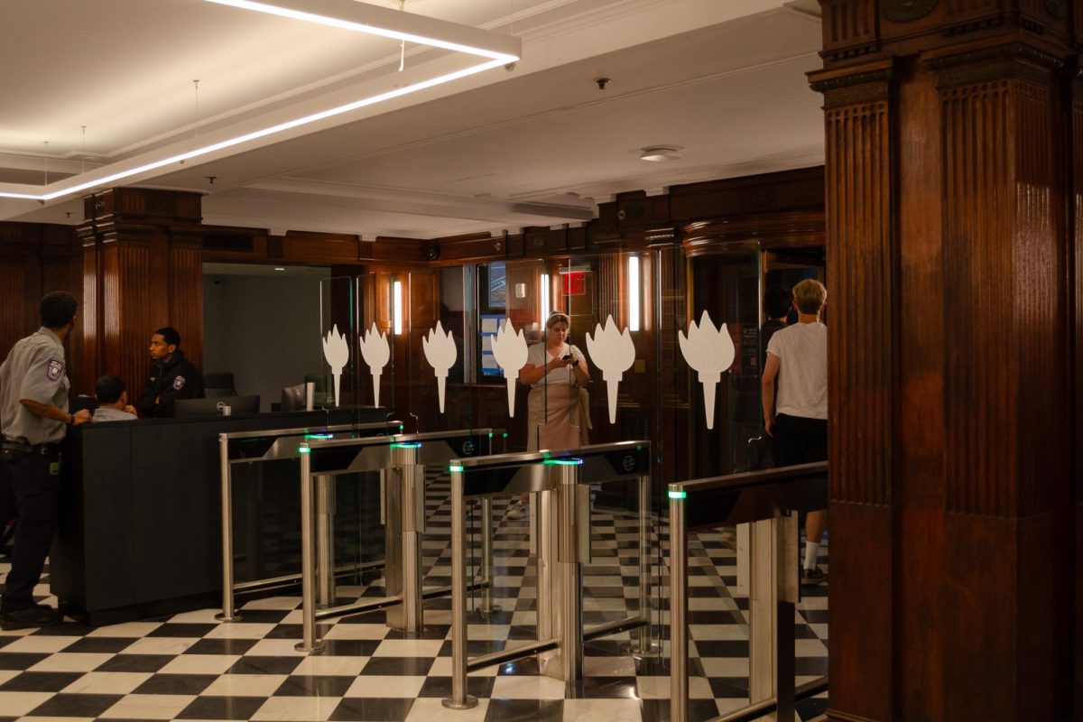 A photograph of a lobby with a black-and-white tile floor, dark wood columns, N.Y.U. security guards and new security entrance gates with the N.Y.U. torch on them.