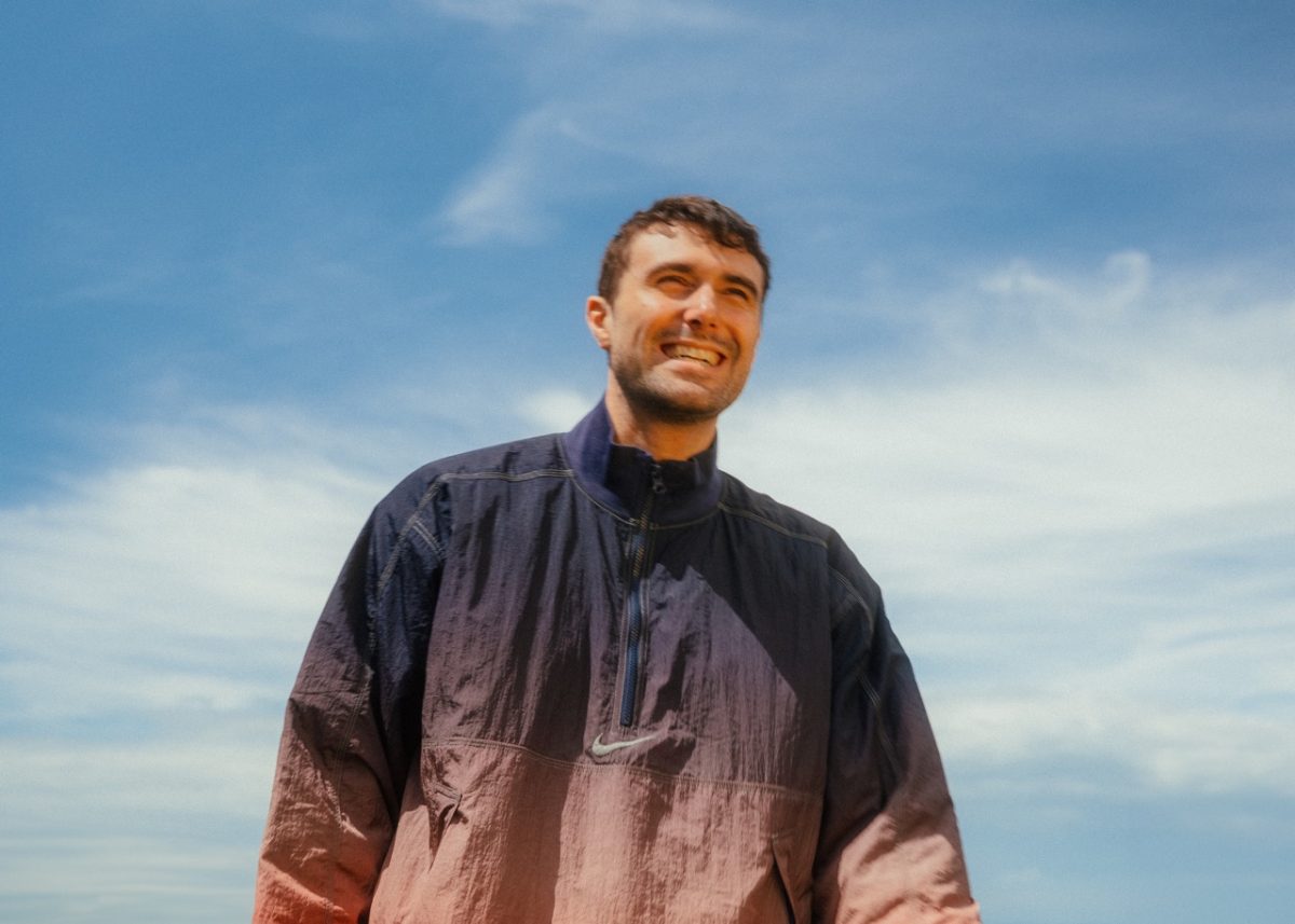 A man wearing a blue zip-up jacket smiles with the sky behind him.