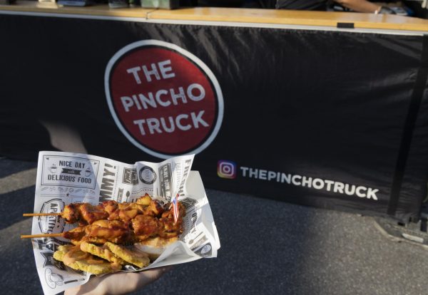 A paper bowl of chicken kabobs with a fried plantains under them, in front of a banner that says “THE PINCHO TRUCK”.