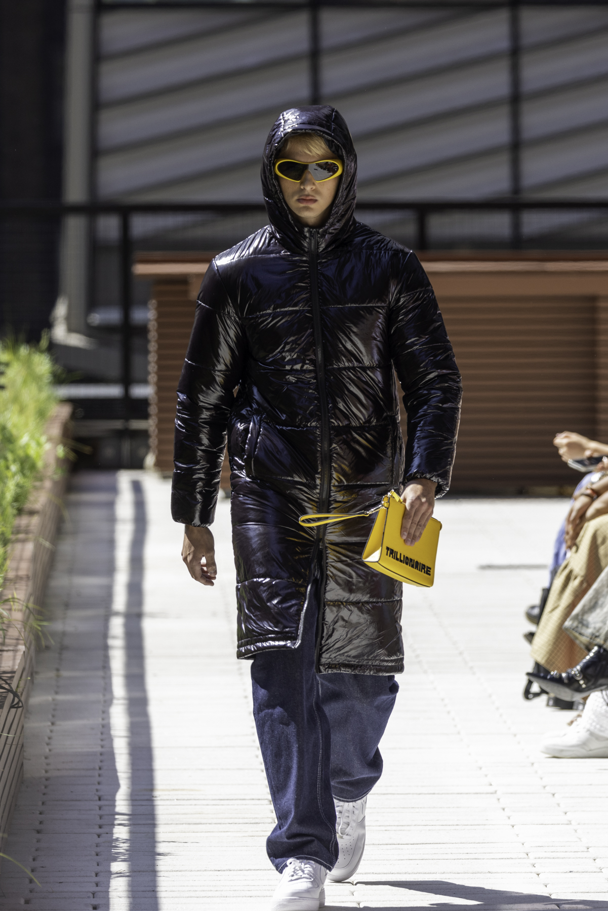 A model wearing a glossy black puffer, white crop top, black glossy pants and white shoes holding a yellow handbag walks down an outdoor runway.