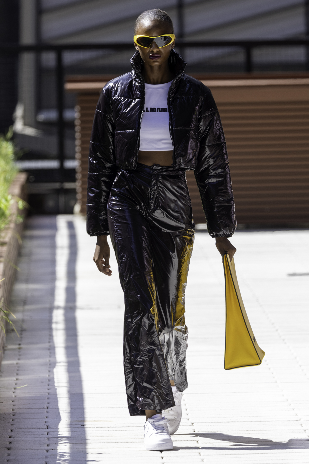 A model wearing a glossy black puffer, white crop top, black glossy pants and white shoes holding a yellow handbag walks down an outdoor runway.