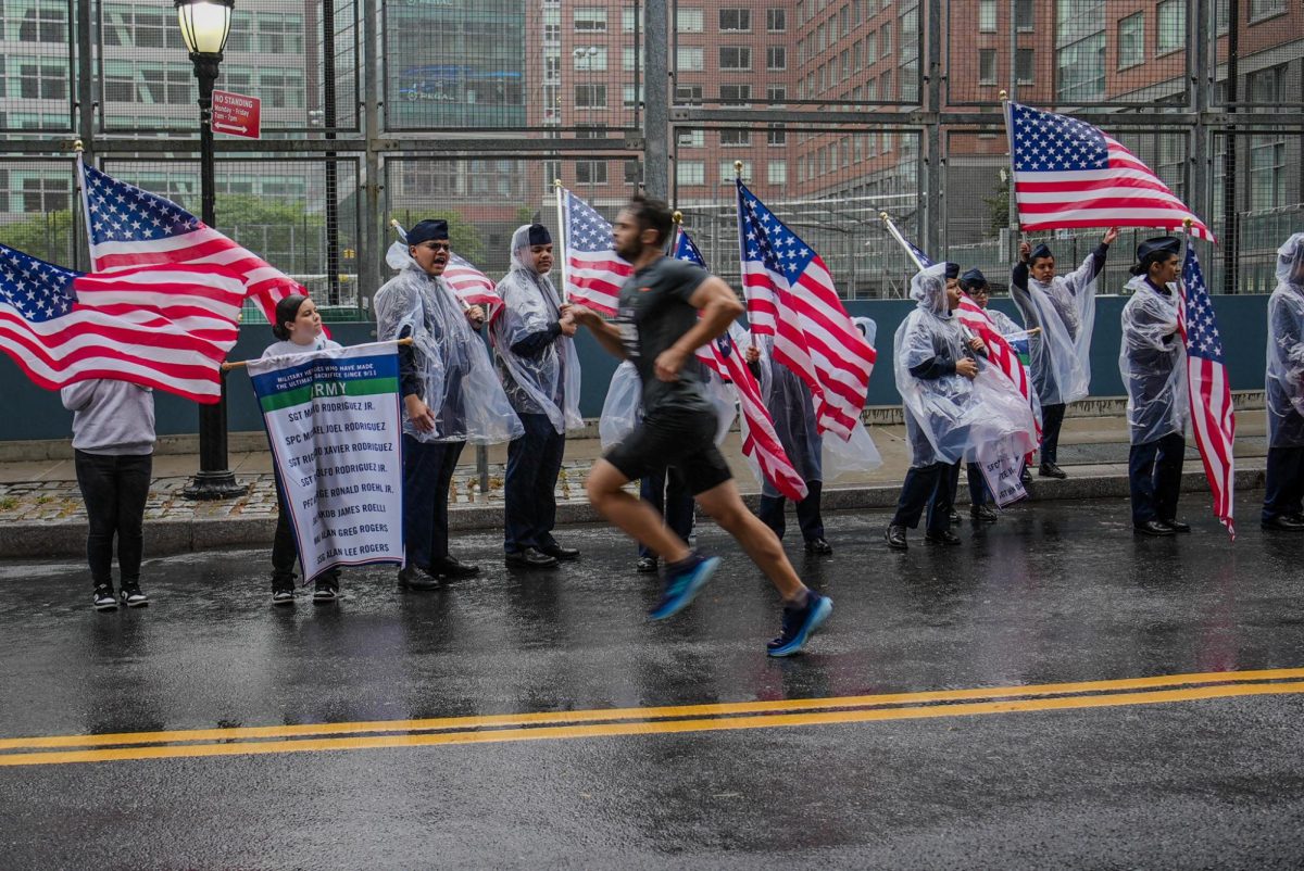 A marathon is happening in the rain with a runner in the foreground. In the background is a group of people in rain ponchos cheering and holding American flags.