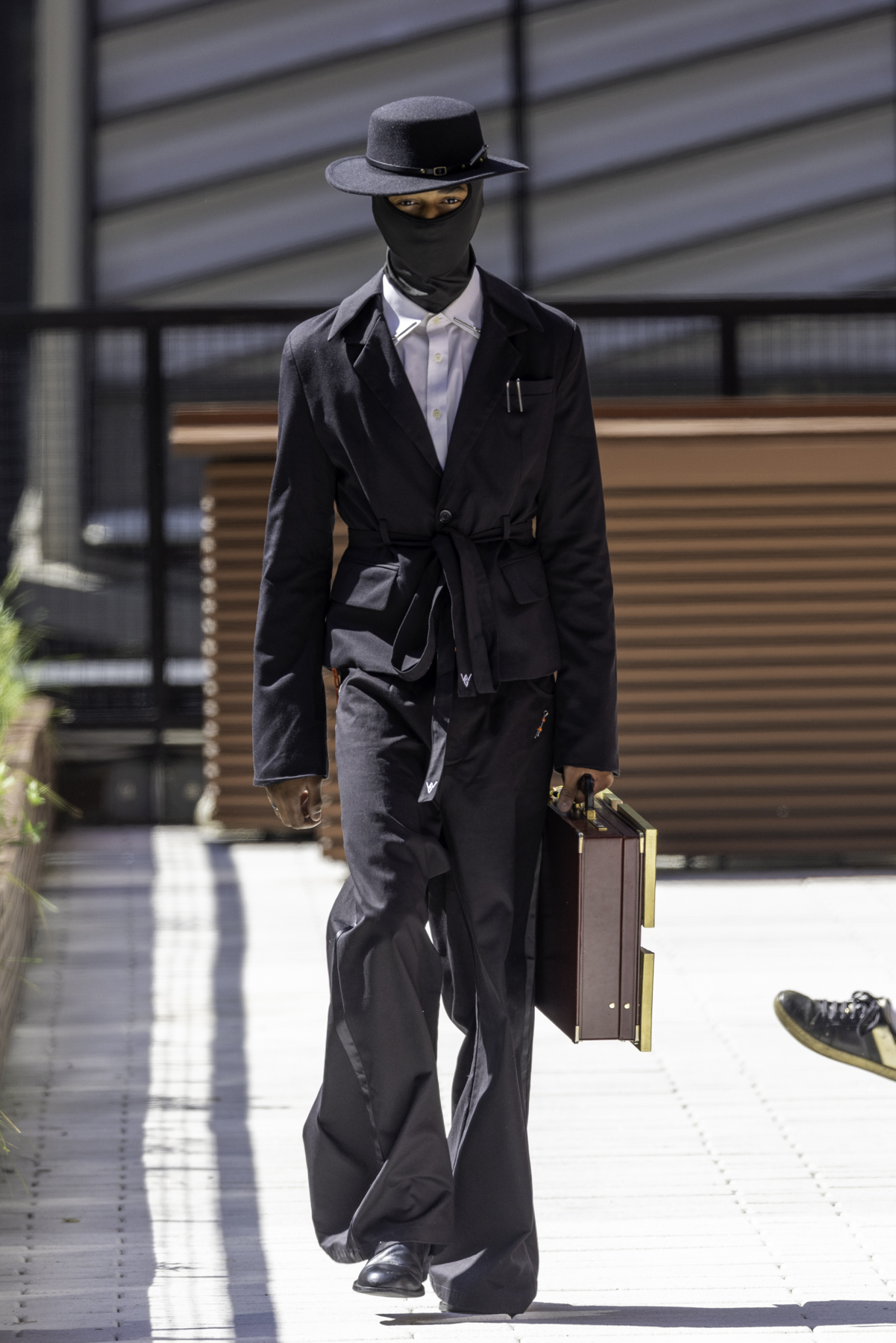 A model wearing a black wide brimmed hat, black ski mask, black suit with a white undershirt, black pants and black dress shoes carrying a brown suitcase walks down the runway.