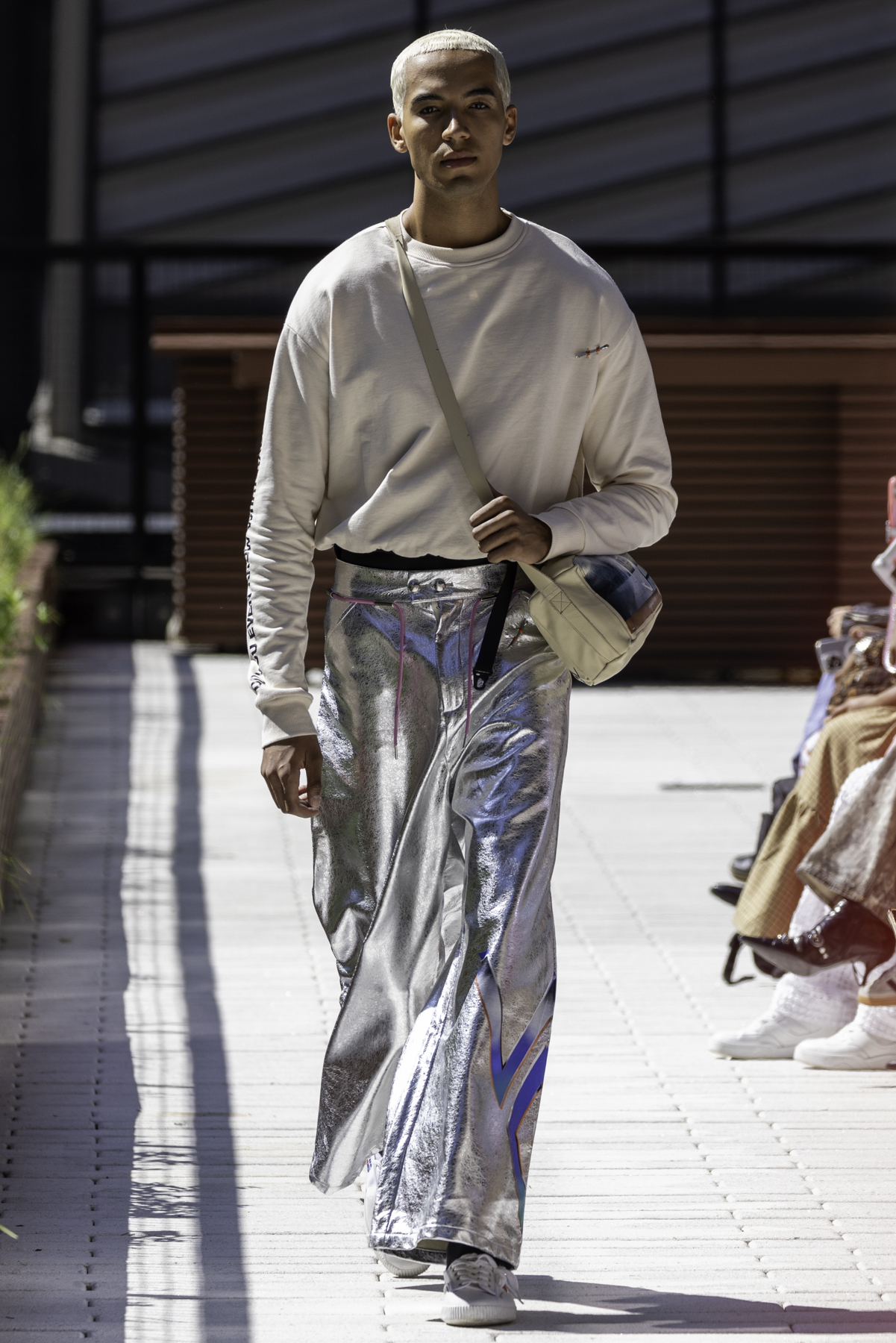 model wearing a beige crewneck sweater, chrome pants, beige sling bag and white shoes walks down the runway.