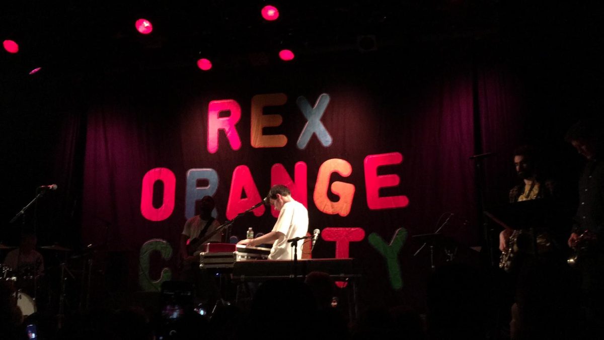 A man is playing the piano on a stage. The words “REX ORANGE COUNTY” are behind him on the stage in colorful letters.