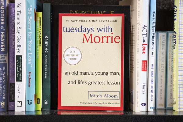 On a bookshelf, a book with "tuesdays with MORRIE" written on it in blue and red and "an old man, a young man, and life's greatest lesson" written in black.