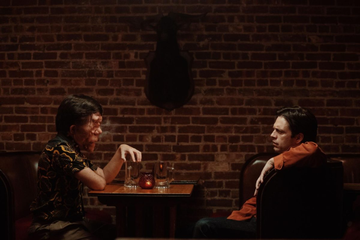 In front of a brick wall, two men sit across from each other at a small table. One man appears normal while the other has a disfigured face.