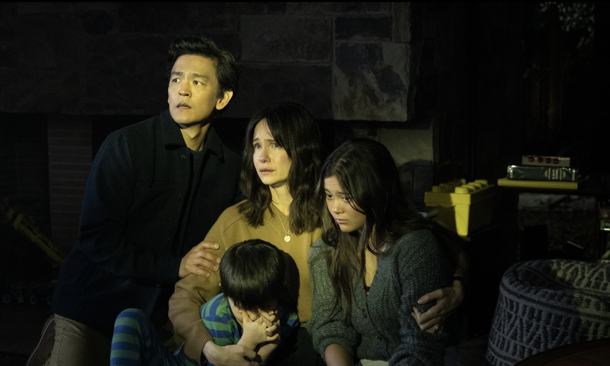 A man in a black shirt, a woman in a brown sweater, a teenage girl in a gray knit sweater and a child in a blue-and-green striped shirt look scared and are holding one another. They are sitting in a dark children’s bedroom.