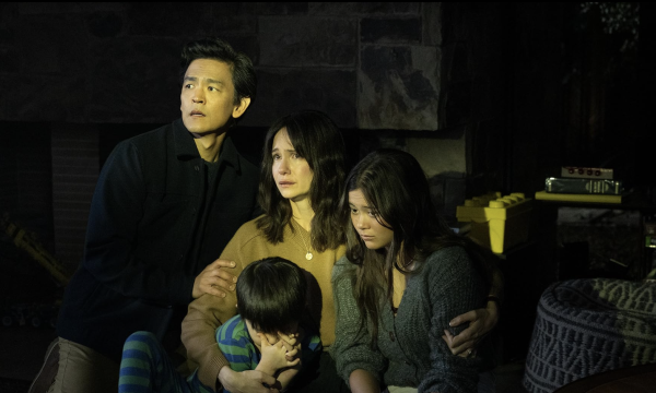 A man in a black shirt, a woman in a brown sweater, a teenage girl in a gray knit sweater and a child in a blue-and-green striped shirt look scared and are holding one another. They are sitting in a dark children’s bedroom.