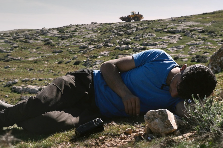 A man in a blue shirt lies on a grassy hill covered in rocks. Behind him is a yellow tractor at the top of the hill.