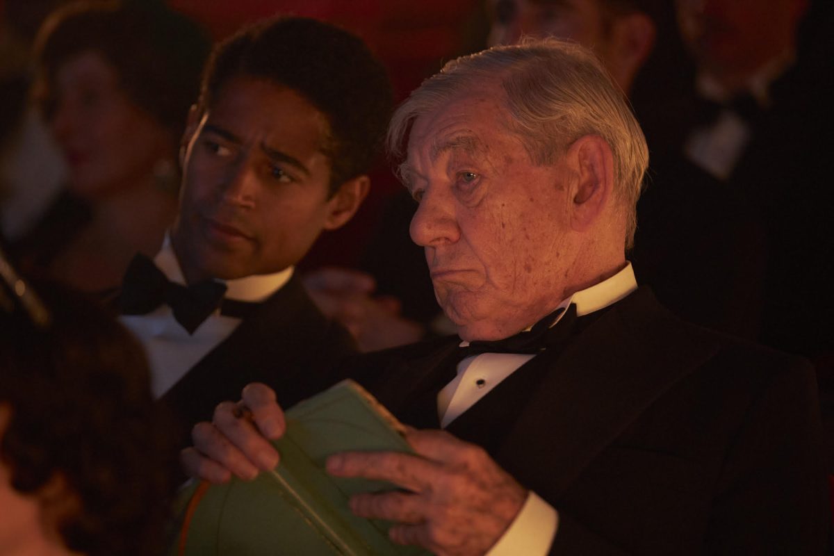 Two men dressed in tuxedos sitting together in an audience. The older man in the foreground is holding a book.