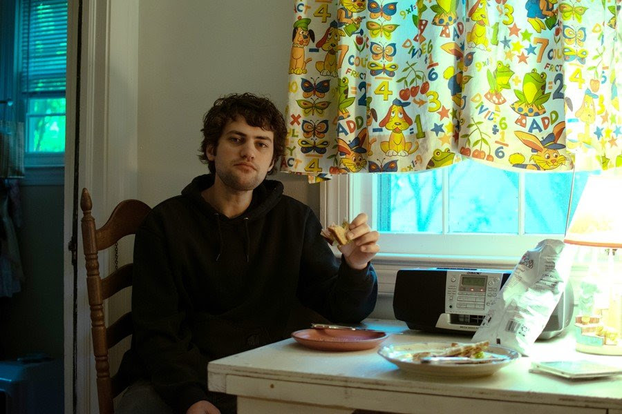 A young man sitting at a kitchen table and eating a sandwich. On the table is a radio and a bag of chips.