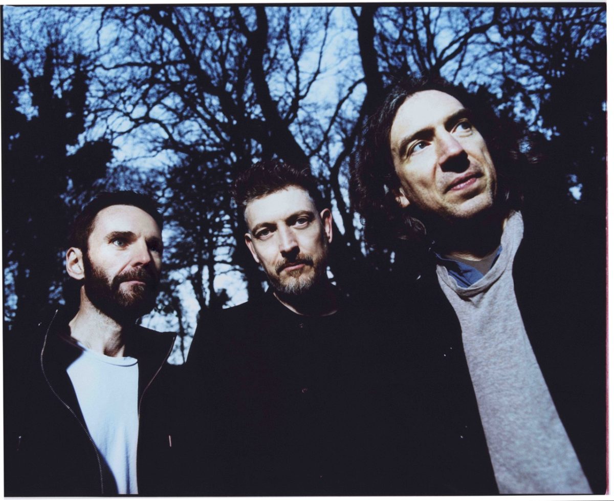 Three men in a forest setting wearing black jackets stand side by side as they look off to the right of the frame.