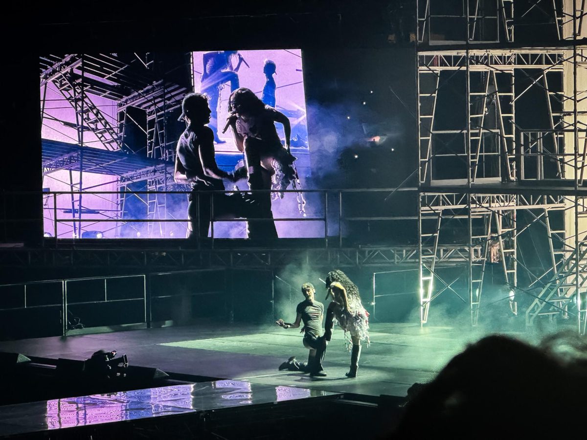 Two singers on stage, one on their knee while the other puts their foot on their leg. In the background, a large jumbotron shows a close-up of the two.