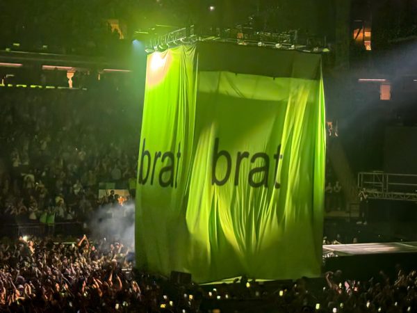 A large crowd of people holding up cell phones pointed at a large neon green curtain that says “BRAT” on it in black arial font.