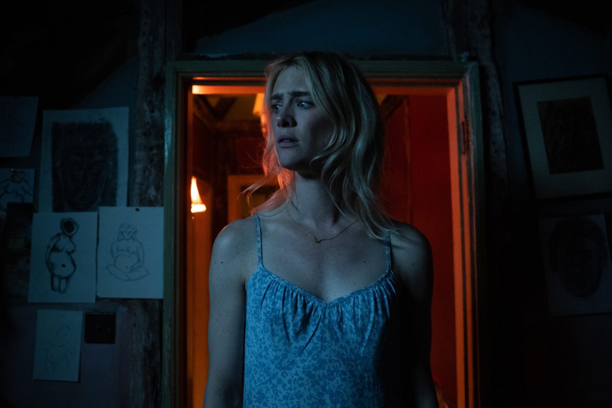 A blonde woman walking into a dark room while looking frightened. Behind her is a doorway with sketches of naked women around it.