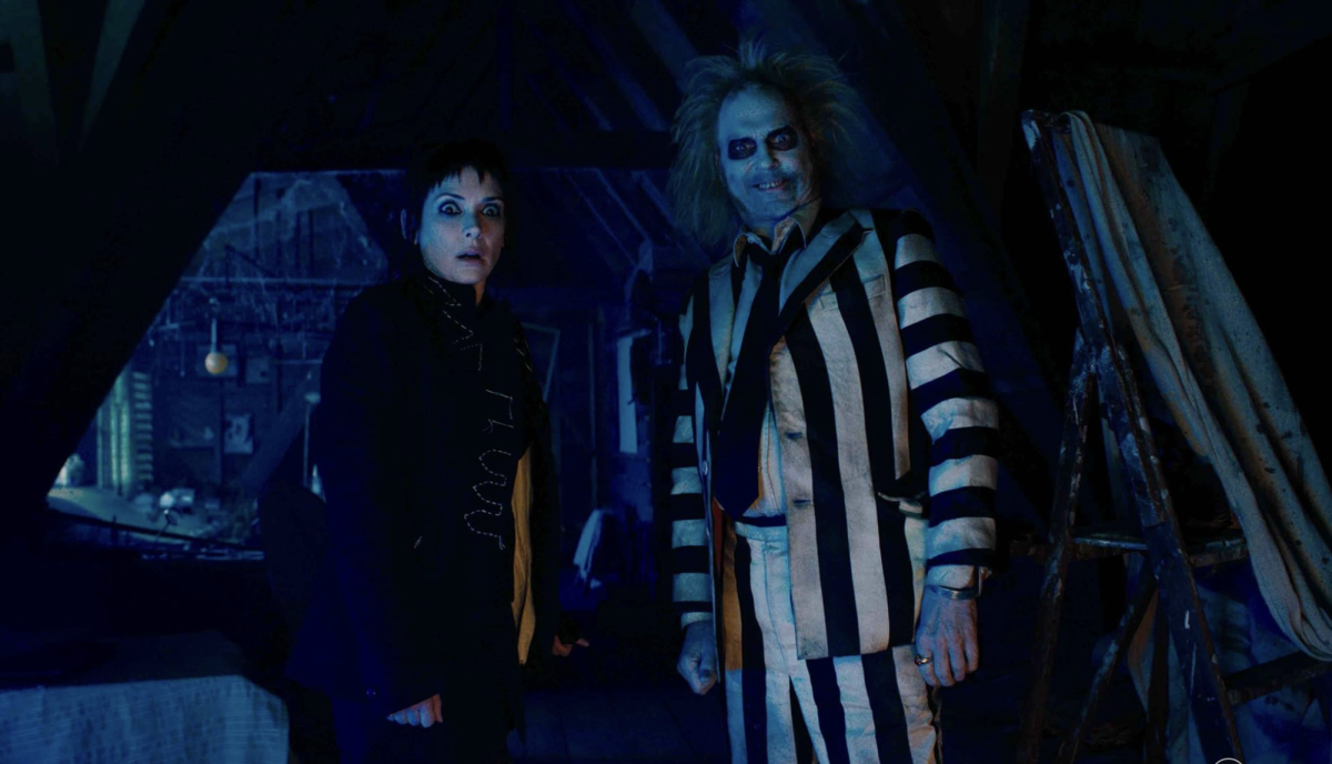 In a dark attic, a woman stands in shock with her eyes wide open while a man next to her in a black and white striped suit and face paint smiles.