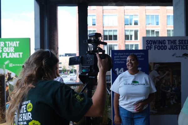 A man interviewing and filming a laughing woman with a “HARLEM GROWN” shirt on.