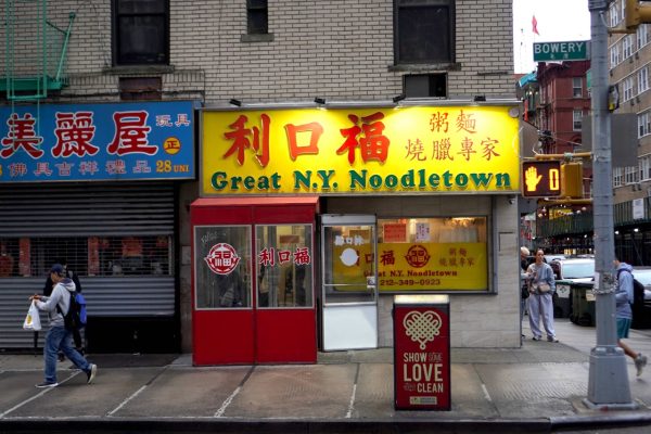 The front of a Chinese restaurant with yellow signs and windows, including red Chinese characters and green letters reading “Great N.Y. Noodletown.”