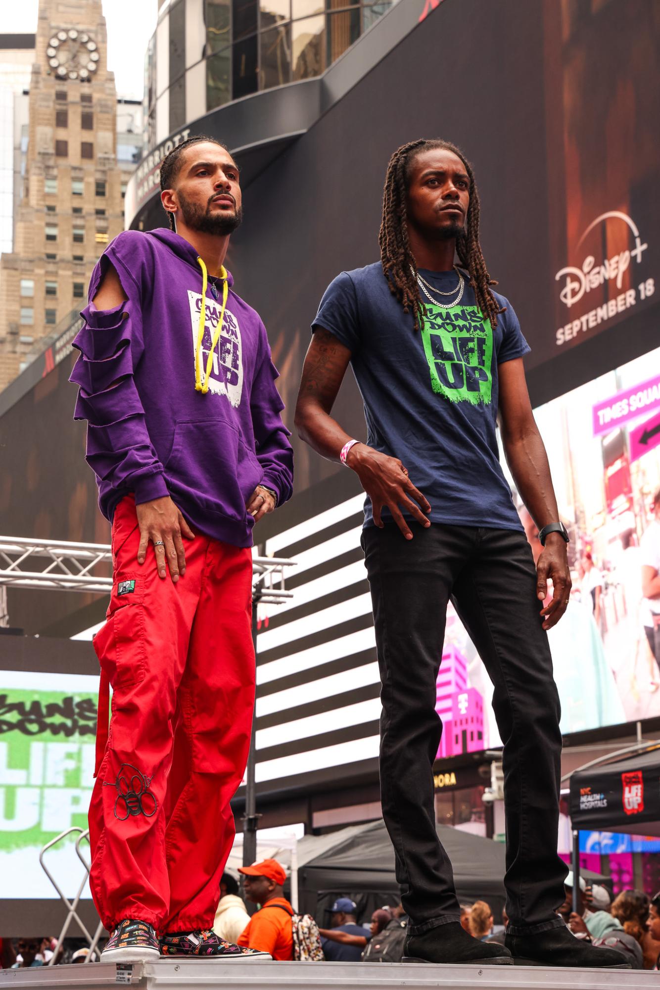 Two models pose next to each other. The one on the left wears a purple hoodie with ripped sleeves and red pants, while the one on the right wears a dark blue shirt and black jeans. Both of their tops say “GUNS DOWN, LIFE UP.”