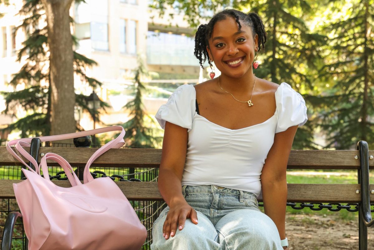 A person in a white top, jeans and pink strawberry earrings sits on a park bench with a pink tote bag.