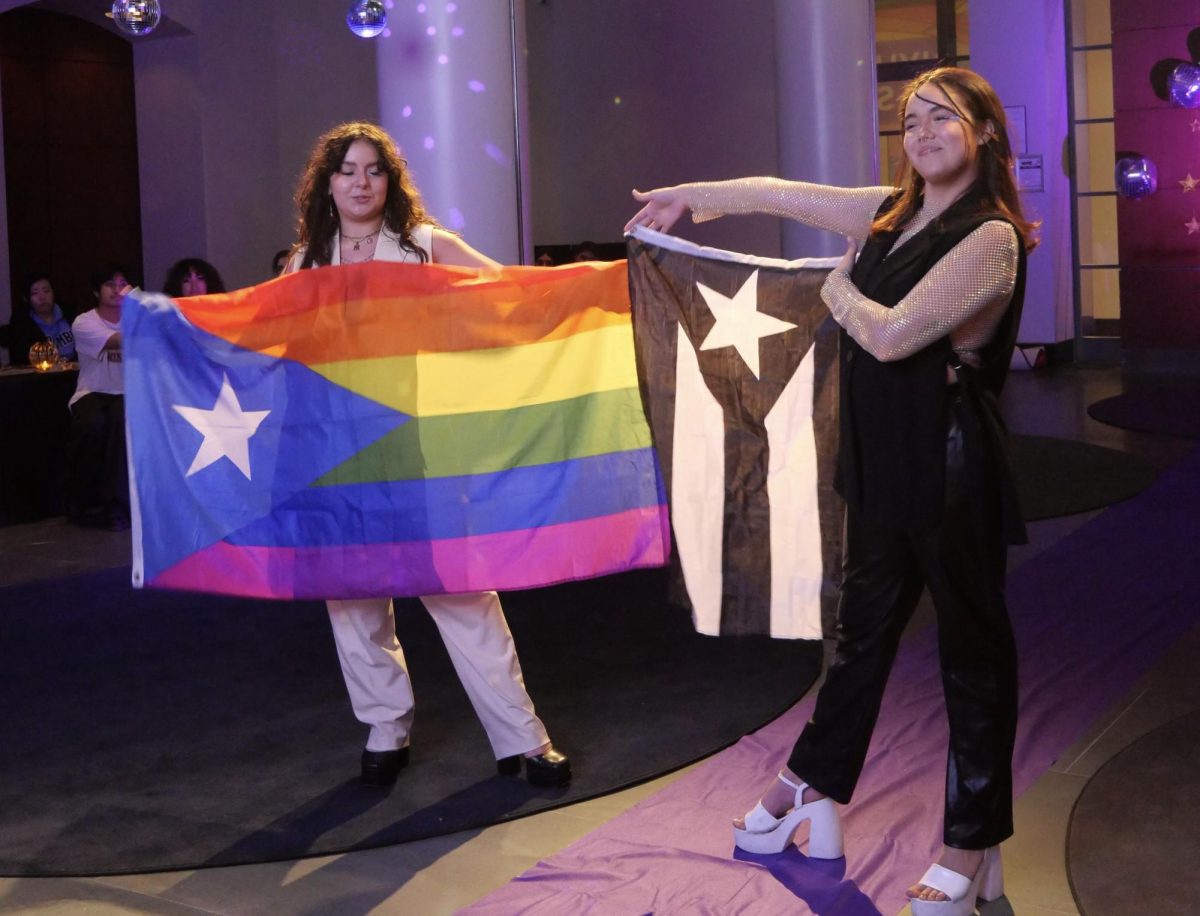 In a purple-lit room, a student holds a striped rainbow flag with a blue triangle and white star while another student next to her holds a black-and-white flag.