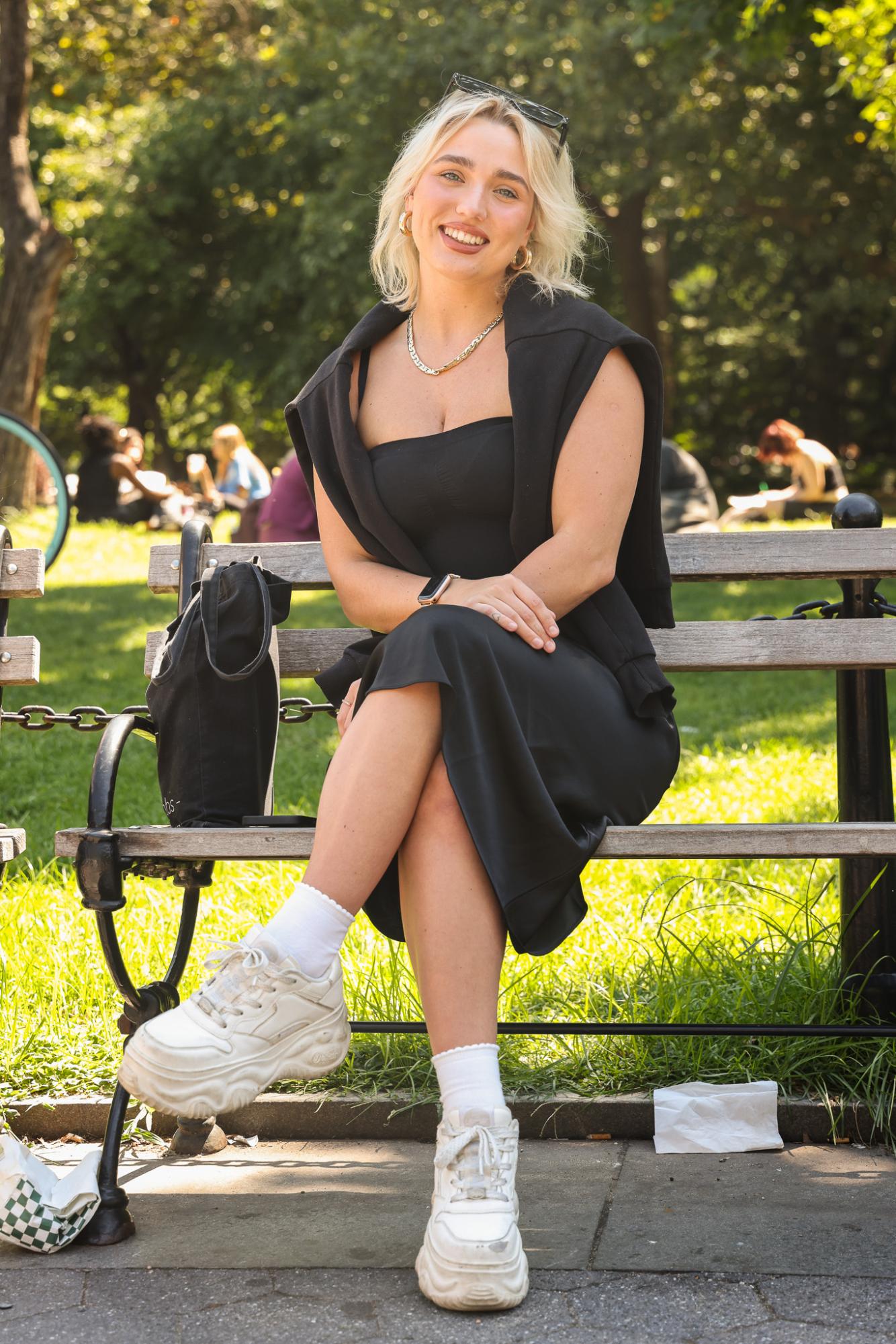 A person sits on a park bench wearing a black dress, white shoes and gold jewelry.