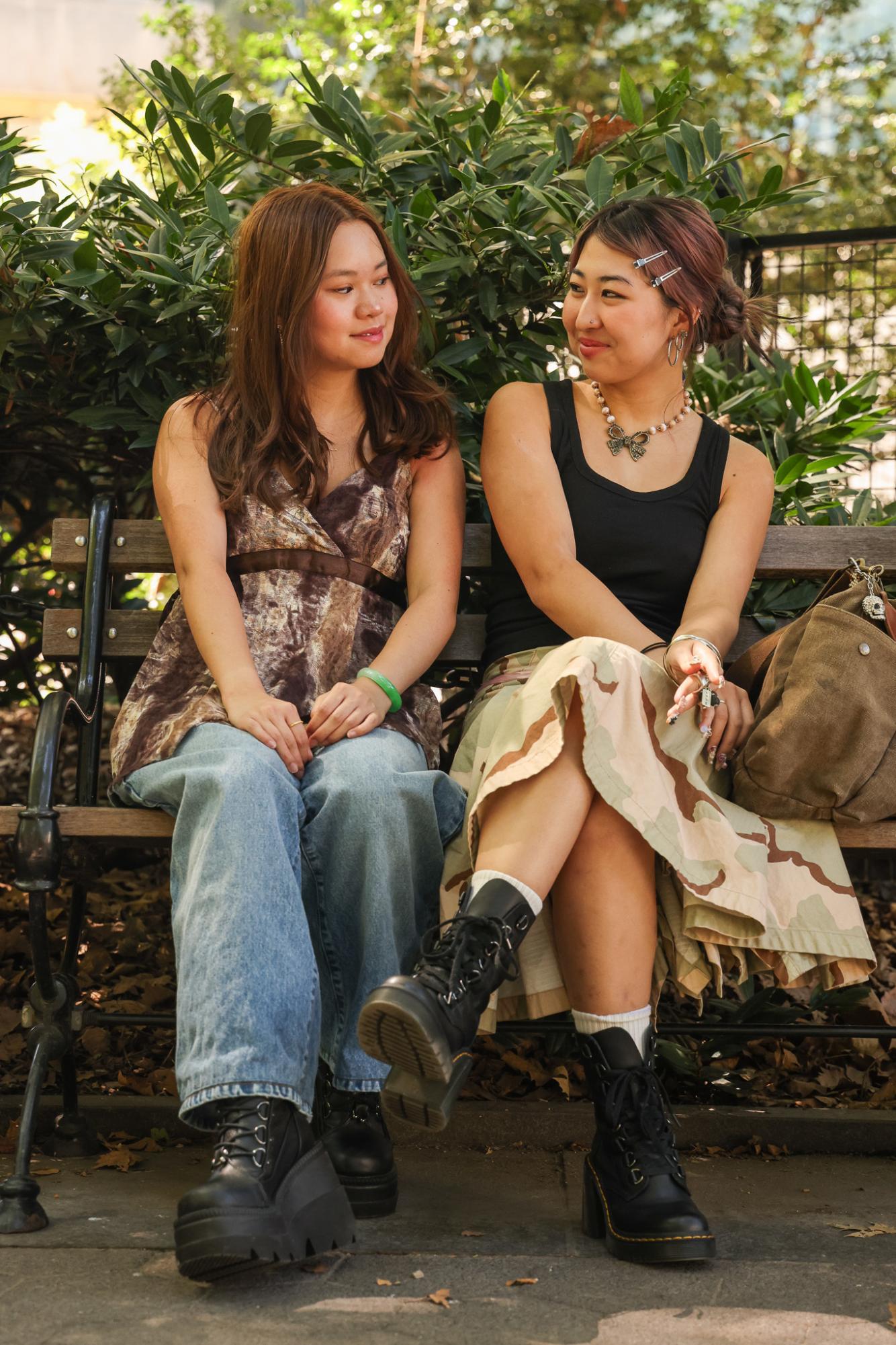 Two people smile at one another while sitting next to each other on a park bench. The one on the left is wearing a brown shirt, blue jeans and black shoes, while the one on the right wears a black top, a camouflage skirt and black shoes.