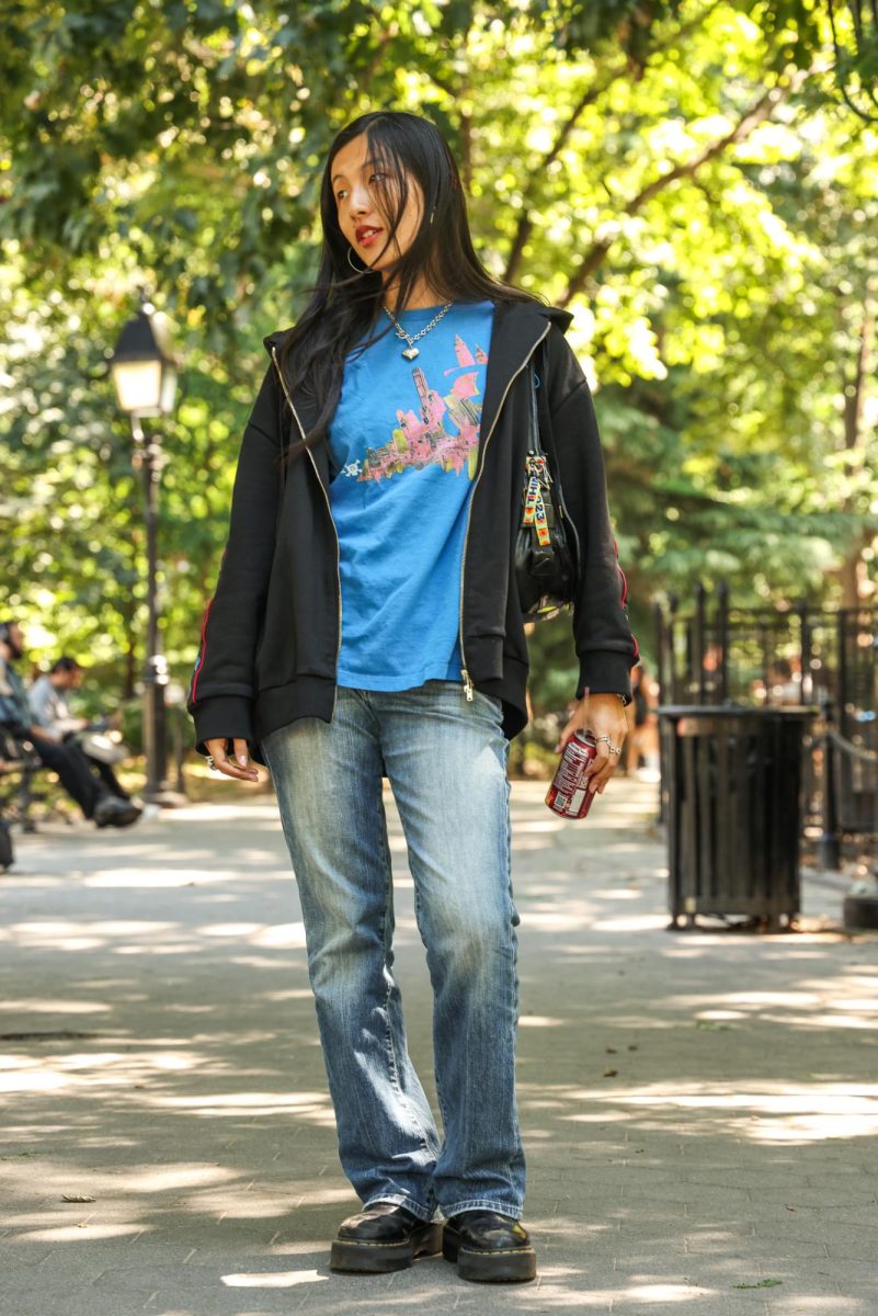 A person poses in a park, standing and looking to the left. They are wearing jeans, a blue shirt and a black zip-up.