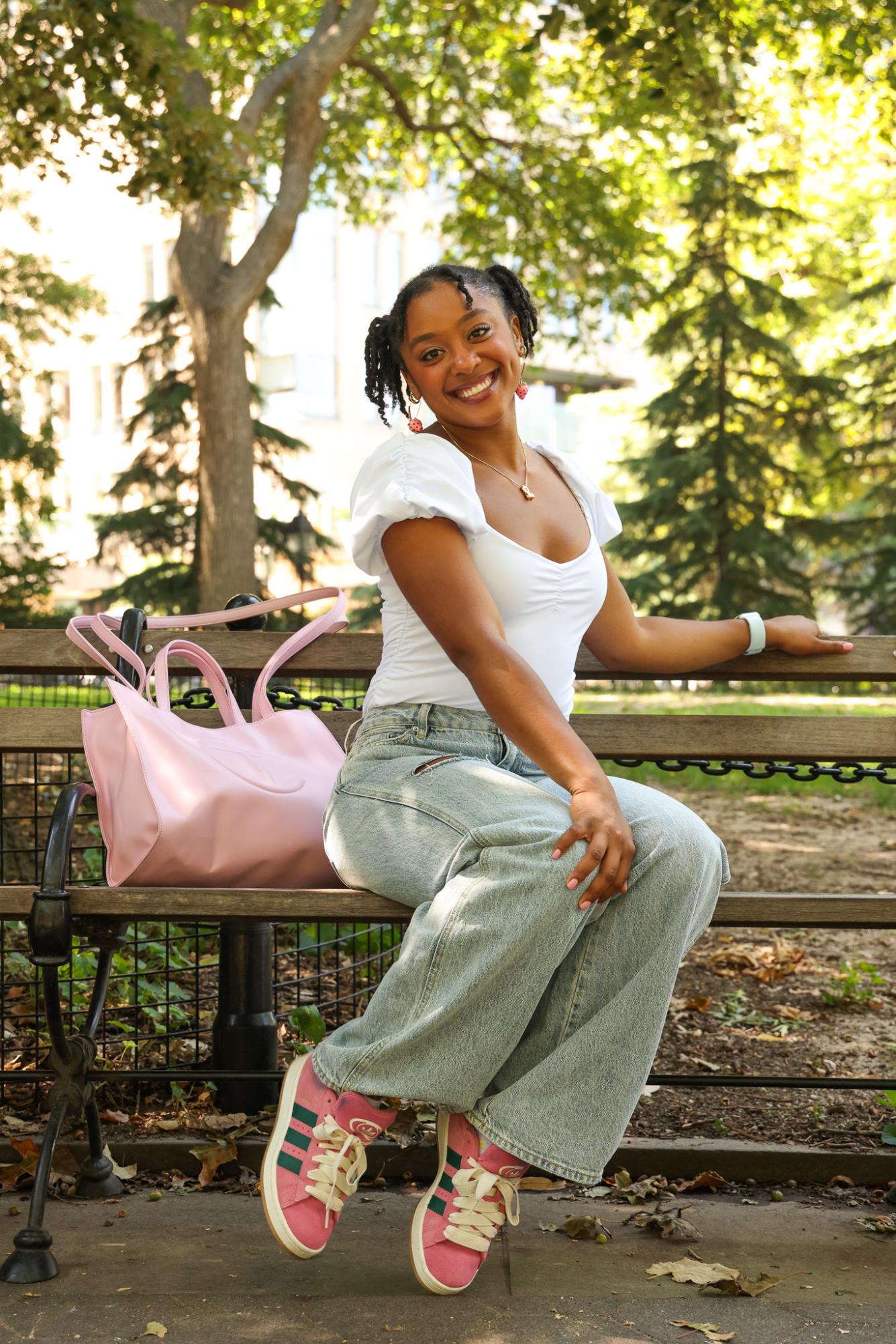 A person in blue jeans and a white shirt sits on a park bench with a pink bag and pink shoes.