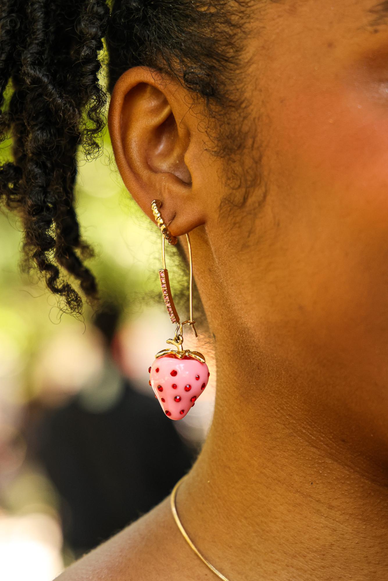 A pink and gold strawberry earring dangles from a person’s ear.