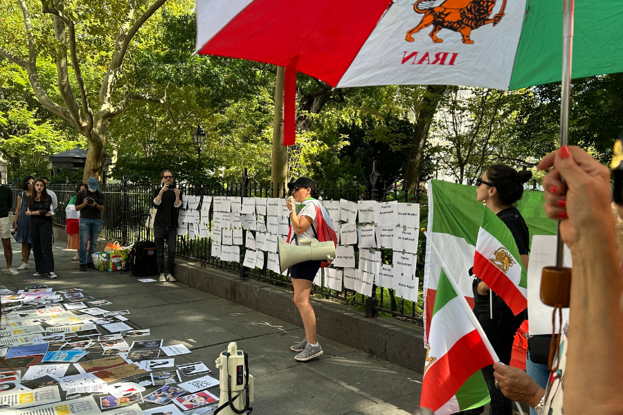 People stand on the sidewalk with a fenced-in park behind them holding Iranian flags and memorabilia. Papers and posters cover the fence and the sidewalk.